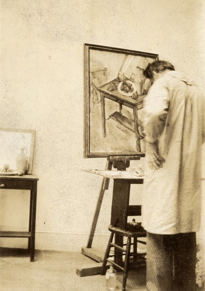 Male artist in a smock seen from behind painting a still life in the style of Paul Cezanne on an easel with artists supplies and objects