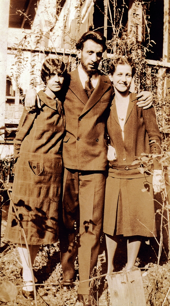 Three people in a yard dressed in formal wear posing, with the man in the middle with his arms wrapped around the women's shoulders