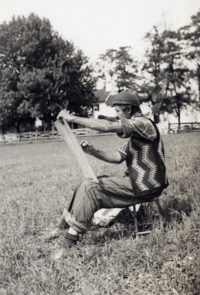 Man wearing a knitted wool vest sitting on a stool in a rural landscape drawing on a drawing board with tress and a farmhouse in the distance