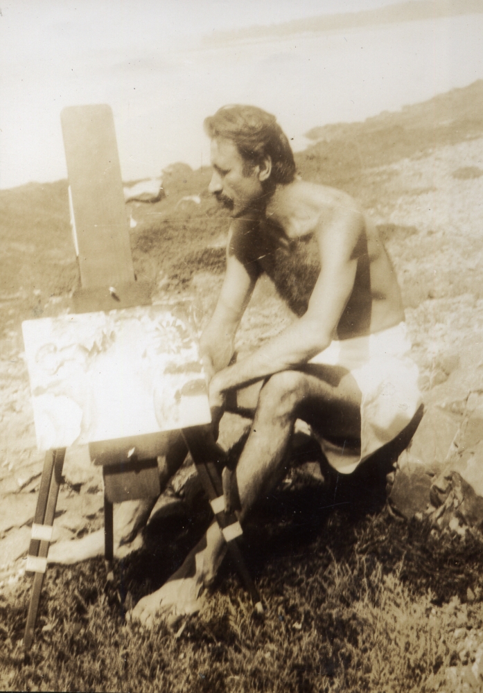 Shirtless man in a bathing suit seated on a rocky beach with a small easel and painting beside him