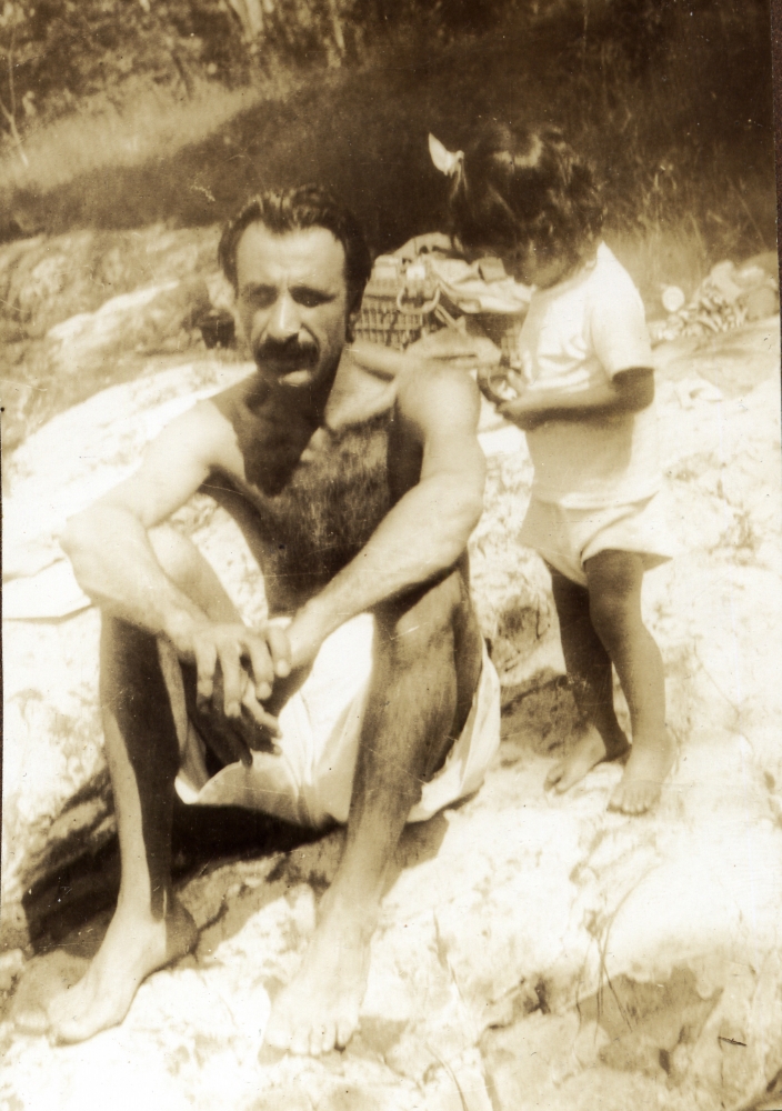 Man with a mustache shirtless in a bathing suit sitting on a rocky beach with a female toddler standing by his left shoulder