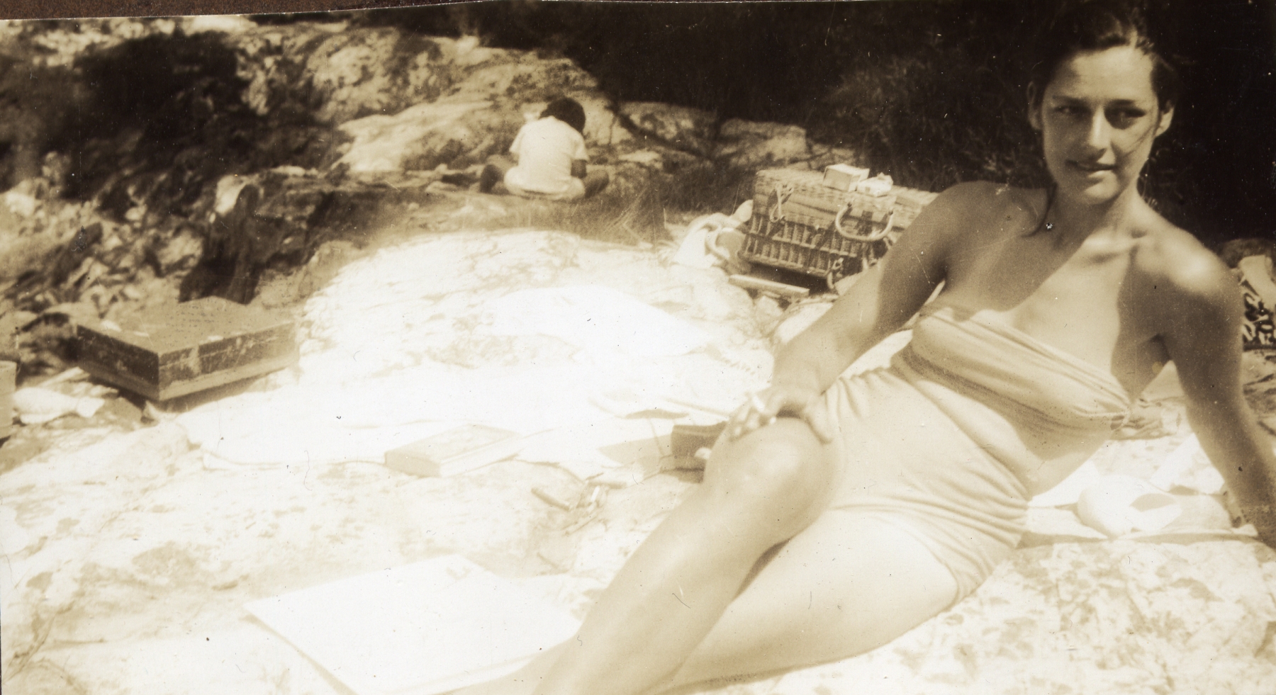 Woman in a bathing suit with her hair wet reclining on a rocky beach, in the distance are a suitcase, wicker basket, and a seated young child seen from behind