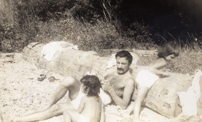 Shirtless man in a bathing suit leaning against a fallen tree trunk on a beach with two young toddlers playing beside him