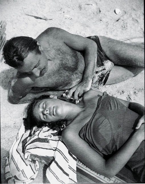 Woman on a beach in a swimsuit sleeping on a towel with a man in a speedo fondly gazing at her