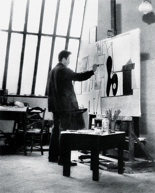 Black-and-white photograph of a man seen from behind in an artist's studio with a large window painting an abstract canvas on an easel