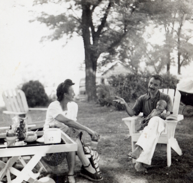 Tea party with a women sitting cross-legged in an Adirondack chair talking to a man sitting in an Adirondack chair, gesturing, and holding an infant