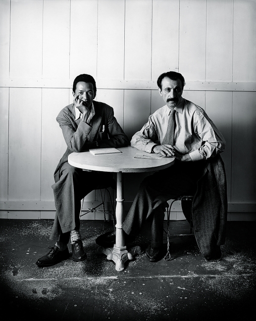 Two men sitting cross-legged in iron chairs leaning on a circular table on which there are papers for drawing
