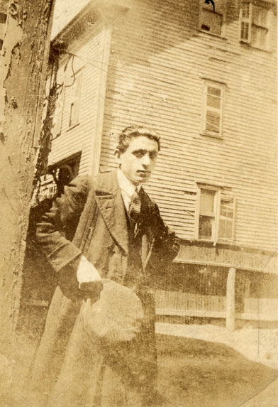 Faded sepia photograph of a young man in a wool jacket holding a wool cap standing before a decrepit house